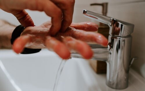 Zwei Hände werden unter fließendem Wasser aus einem modernen, silbernen Wasserhahn gehalten. Es scheint, als würde sich die Person die Hände waschen. Der Fokus liegt auf den Händen und dem Wasserhahn, der Hintergrund ist unscharf. | © Claudio Schwarz (Unsplashed)