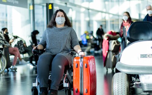 Frau im Eingangsbereich des Flughafens sitzt in einem Elektrorollstuhl mit kleinem roten Koffer in der linken Hand | © Help-24 GmbH, ackerblau