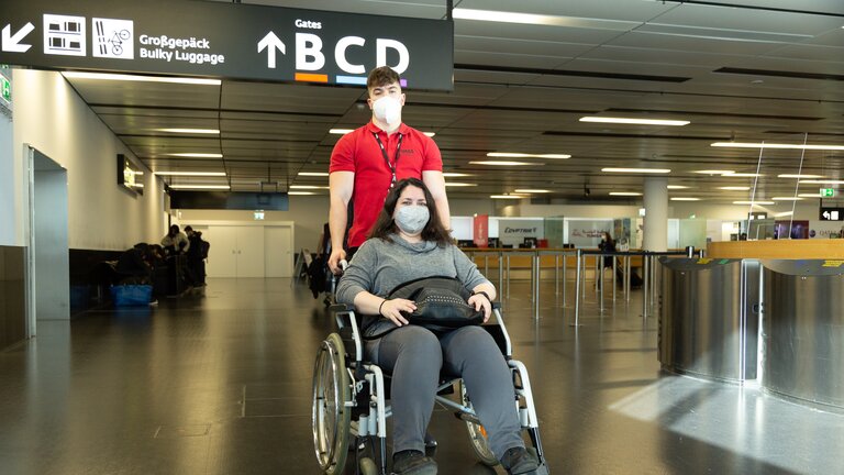 Am Flughafen: Frau sitzt in einem Rollstuhl mit Handtasche auf dem Schoß und wird von einem Mitarbeiter geschoben. | © Help-24 GmbH, ackerblau