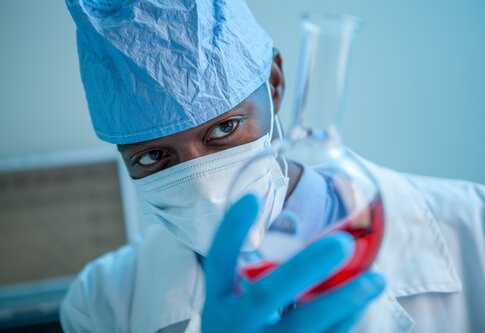 Foto von einem Mann, der Arztkleidung und eine medizinische Maske trägt, in seiner Hand hält er ein Reagenzglas, das mit einer roten Flüssigkeit gefüllt ist. | © Unsplash