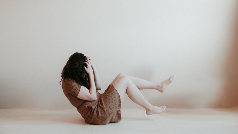 Das Bild zeigt eine Frau mit langen schwarzen Haaren in einem braunen Kleid, die auf dem Boden vor einer weißen Wand sitzt. Sie hält die Hände vor ihr Gesicht und hat die Füße leicht angewinkelt. | © Unsplash