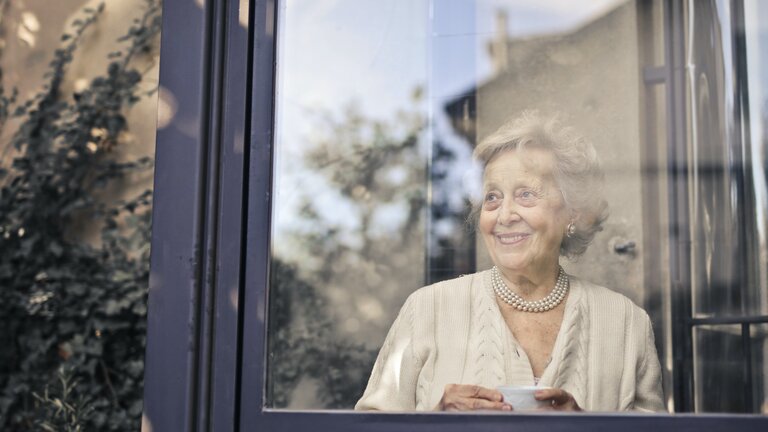 Eine ältere Frau guckt aus einem Fenster und lächelt. | © pexels