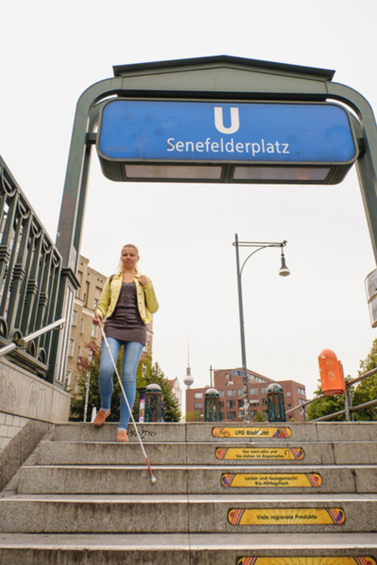 Das Bild zeigt eine junge Frau, die eine Treppe an einer U-Bahn-Station heruntergeht. Die Station heißt "Senefelderplatz", was auf dem blauen Schild über dem Eingang steht. Die Frau hat blonde Haare, trägt eine gelbe Jacke, ein dunkles Oberteil und blaue Jeans. Sie benutzt einen weißen Blindenstock. Im Hintergrund sind einige Gebäude und der Berliner Fernsehturm sichtbar. | © Andi Weiland / Gesellschaftsbilder