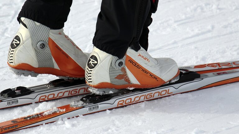 Man sieht die Füße, eines Ski-Langläufer. Die Farben der Schuhe und der Skier sind orange und weiß. Der Boden ist mit Schnee bedeckt. | © Pixabay.com