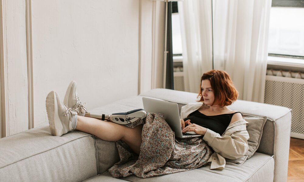 Eine Frau mit einer Beinprothese sitzt bequem auf einem Sofa und surft mit ihrem Laptop im Internet oder besucht unsere Community. | © Pexels / Vlada Karpovich