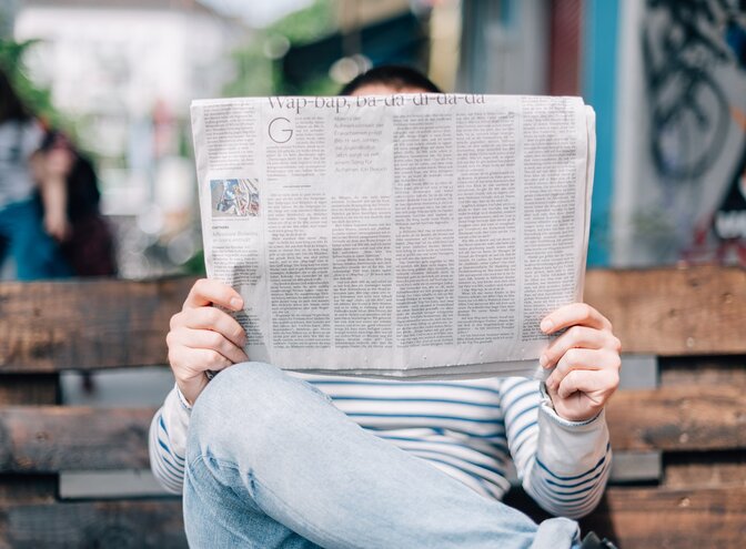 Ein Mann sitzt auf der Bank und liest die Zeitung so, dass man sein Gesicht nicht sieht. | © Unsplash / Roman Kraft