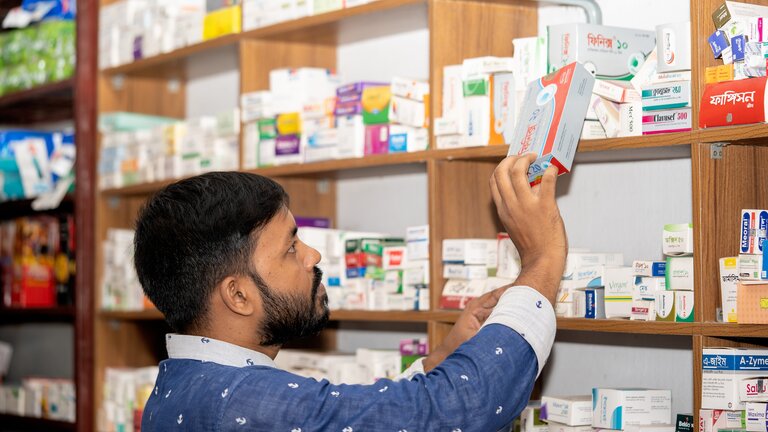Foto eines Apothekers, der gerade eine Schachtel mit Medikamenten aus einem Regal nimmt.