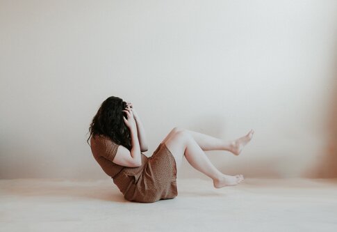 Eine Frau in einem braunen Kleid und mit dunklen Locken sitzt in einem weißen Raum und hält ihre Hände an den Kopf. | © Priscilla du Preez/unsplash