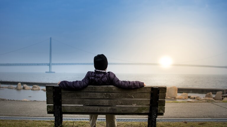 Eine Person sitzt auf einer Bank am Ufer und beobachtet den Sonnenuntergang. | © Sid Leigh/unsplash