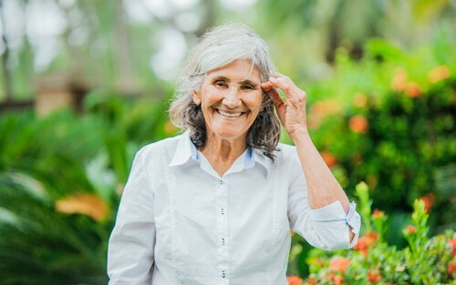 Eine Seniorin mit weißer Bluse steht lächelnd in einem Garten. | © pexels