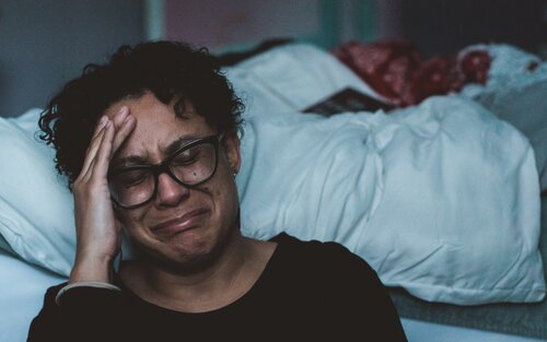 Eine verzweifelte Frau sitzt neben ihrem Bett und hält sich die Hand an die Stirn während sie weint. | © Claudia Wolff/unsplash