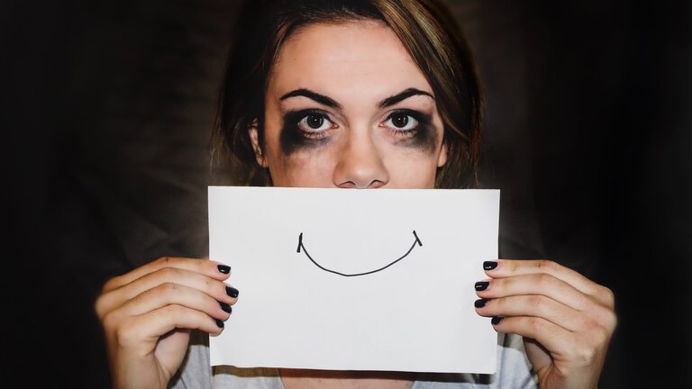 Weinende Frau, deren Mascara stark verwischt ist, hält sich ein Blatt Papier vor den Mund auf dem ein Lächeln gezeichnet ist. | © Sydney Sims/unsplash