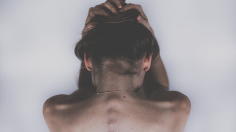 Eine Frau mit nacktem Oberkörper wird von hinten fotografiert. Sie befindet sich in einem weißen Raum und hält ihren Kopf in die Hände. | © pixabay