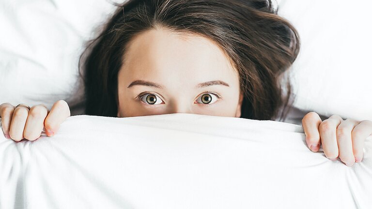 Eine Fraumit braunen Haaren und grünen Augen liegt im Bett und hat ihr halbes Gesicht unter der Bettdecke versteckt. | © unsplash