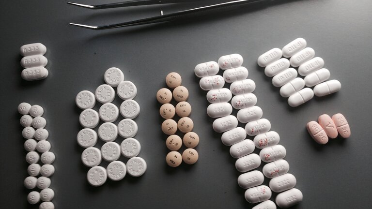 Tabletten in verschiedenen Größen und Formen ligen sortiert in sieben Gruppen auf einer grauen Fläche. über den Medikamenten liegt eine große silberfarbene Pinzette.  | © unsplash