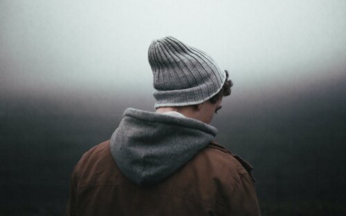 Ein junger Mann wird von hinten fotografiert und schaut dabei leicht zu Boden. Er trägt eine graue Mütze und eine braune Jacke, aus der eine graue Kapzue hervorschaut.  | © unsplash