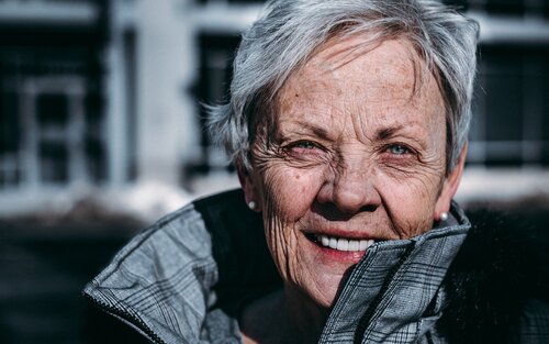 Nahaufnahme des Gesichts einer älteren Dame mit kurzen grauen Haaren und blauen Augen. Sie trägt eine graue Jacke mit großem Kragen und Perlenohrringe. Sie lächelt in die Kamera. | © unsplash