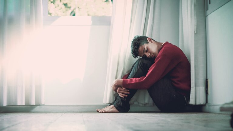 Ein deprimierter Mann sitzt unter einem Fenster auf dem Boden und umklammert dabei seine Beine. Seinen Kopf lehnt er dabei auf die Knie und hat seine Augen geschlossen. | © unsplash