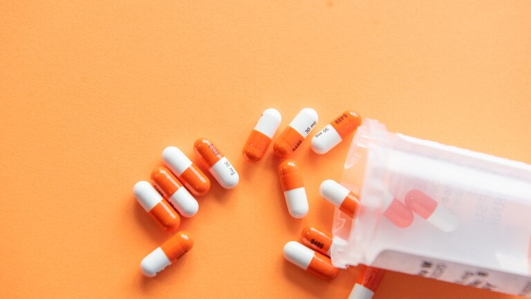 weiß-orangene Pillen liegen ausgeschüttet aus einem Behälter auf einer orangenen Fläche. | © unsplash