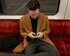 Ein Mann mit einer Brille, grauen Hose und braunem Mantel sitzt mit einem Smartphone in der Hand im Zug. | © Mart Production/ pexels.com