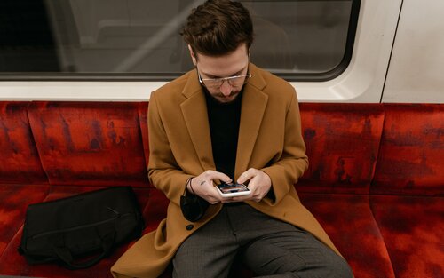 Ein Mann mit einer Brille, grauen Hose und braunem Mantel sitzt mit einem Smartphone in der Hand im Zug. | © Mart Production/ pexels.com