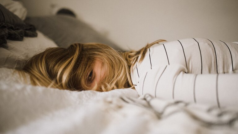 Oberkörper einer Frau müde auf dem Bett liegend. Das Gesicht ist mit Haaren bedeckt | © unsplash