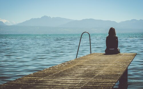 Foto von einer Frau, die auf einem Steg sitzt und aufs Wasser blickt. | © Unsplash
