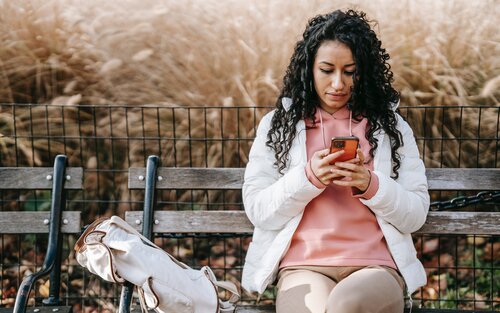 Eine Frau mit schwarzen Locken und einer weißen Jacke sitzt auf einer Holzbank und blickt auf das Smartphone in ihrer Hand | © pexels