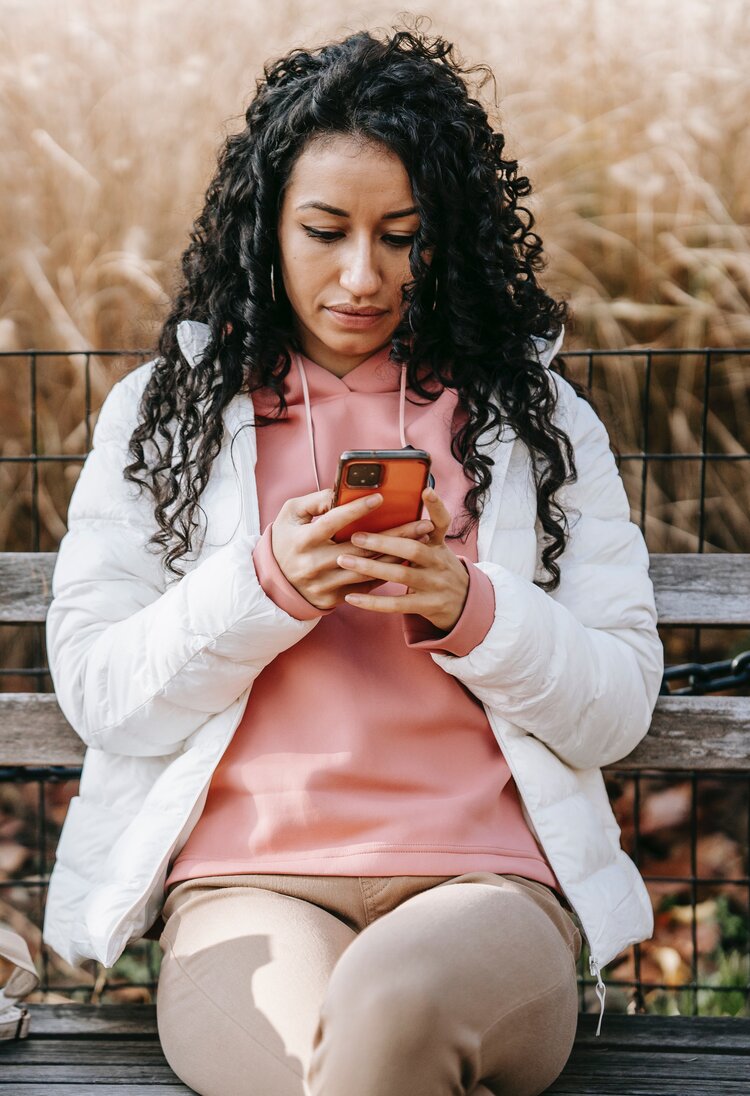 Eine Frau mit schwarzen Locken und einer weißen Jacke sitzt auf einer Holzbank und blickt auf das Smartphone in ihrer Hand | © pexels