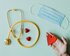 Auf einem hellplauen Tisch liegt ein Stethoskop, eine medizinische Maske und zwei Schokoladenherzen, die in rot-glänzendem Papier verpackt sind. Eine Hand, in der ein rotes Papierherz liegt, ragt in das Bild. | © Karolina Grabowska / Pexels.com