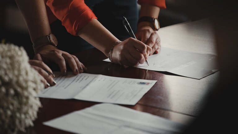Man sieht die Hände von zwei Personen, die grade Dokumente, die auf einem Tisch liegen unterschreiben. | © Romain Dancre/ unsplash