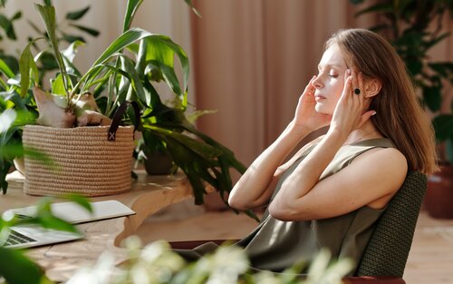 Eine blonde Frau sitzt am Schreibtisch, von Grünpflanzen umgeben, und hält beide Hände an ihren Kopf. Die Augen sind geschlossen. | © Mikael Blomkvist / Pexels