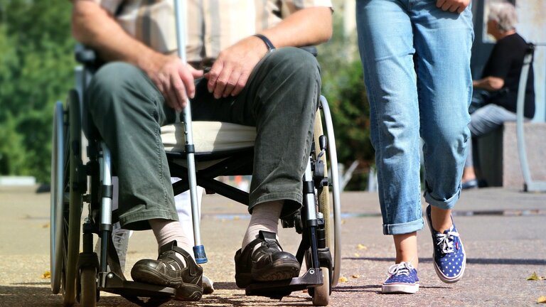 Nahaufnahme von einem Mann in Rollstuhl, der auch eine Krücke in der Hand hält. Eine Frau läuft nebenher.  | © pixabay