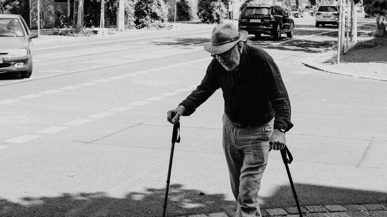 Ein schwarz-weißes Bild von einem älteren Mann, der mithilfe von zwei Stöcken auf der Straße geht. | © unsplash