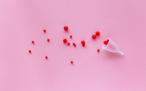 Das Bild zeigt einen rosa Hintergrund mit mehreren kleinen roten Pompons, die verstreut sind. In der rechten unteren Ecke befindet sich eine Menstruationstasse, die umgedreht ist und einige der roten Pompons enthält. | © Nataliya Vaitkevich (Pexels)