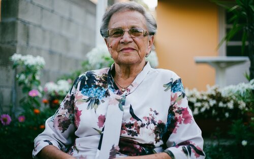 Portrait einer älteren Dame mit Brille und geblümter Bluse vor einer Hauswand. | © Damir Bosnjak/unsplash