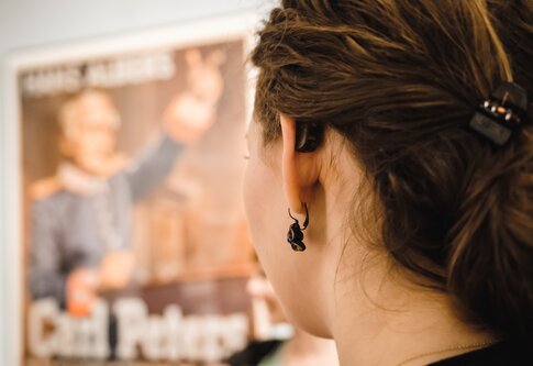 junge Frau von hinten mit Hörgerät sieht auf ein verschwommenes Plakat | © Andi Weiland/ Gesellschaftsbilder.de