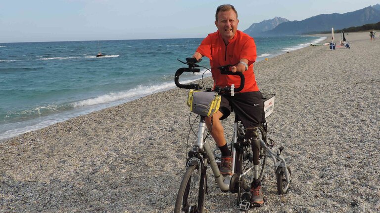 Ein Mann ohne Hände und einer Beinprothese am linken Bein sitzt auf einem Pedelec mit Stützradsystem am Strand. Der Mann trägt eine schwarze kurze Sporthose und ein rotes T-Shirt. | © Ludwig Lübbers