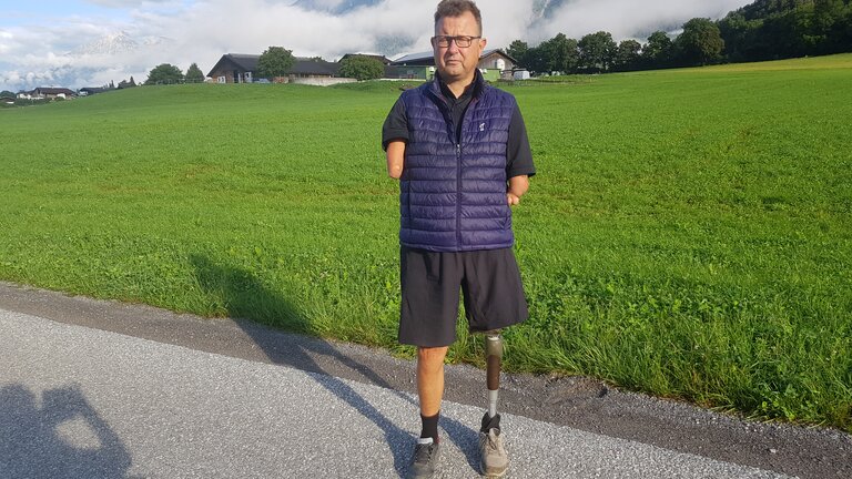 Ein Mann ohne Hände mit einer Beinprothese am linken Bein steht vor einer grünen Wiese. Im Hintergrund sind Berge, Bäume und Wolken. Er trägt eine kurze schwarze Sporthose und eine blaue Weste. | © Ludwig Lübbers