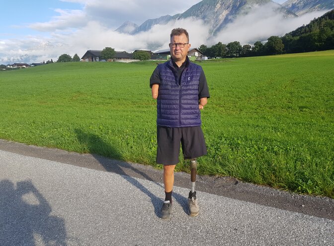 Ein Mann ohne Hände mit einer Beinprothese am linken Bein steht vor einer grünen Wiese. Im Hintergrund sind Berge, Bäume und Wolken. Er trägt eine kurze schwarze Sporthose und eine blaue Weste. | © Ludwig Lübbers