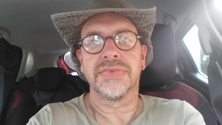 Das Foto zeigt einen mittelalten Mann, der in einem Auto sitzt. Er trägt ein olivgrünes T-Shirt, eine Brille mit runden Brillengläsern und einem schwarzen Rahmen sowie einen grauen Sonnenhut. Er hat Bartstoppeln und guckt freundlich in die Kamera. | © Tobias N. privat