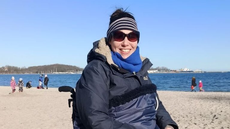 Wiebke im Rollstuhl am Strand mit einer Decke, Stirnband und Sonnenbrille | © Wiebke