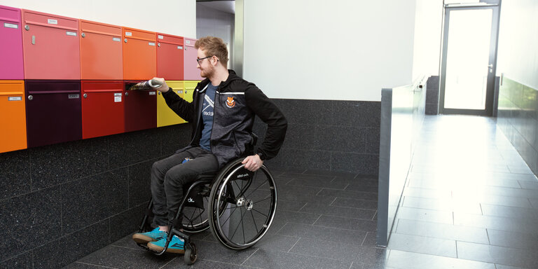 Ein junger Mann im Rollstuhl im Eingangsbereich des Hauses, in dem sich seine rollstuhlgerechte Wohnung befindet | © © adira.de, Fotograf: Daniel George