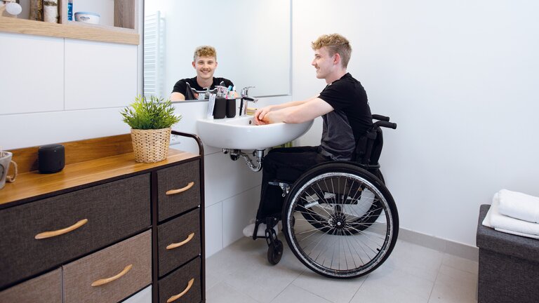 Ein junger Mann im Rollstuhl sitzt an seinem rollstuhlgerechten Waschtisch im Badezimmer und wäscht sich die Hände. Er lächelt den Betrachter über den Spiegel an. | © ©adira.de, Fotograf: Daniel George