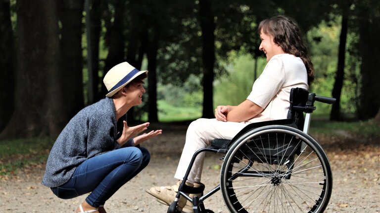 Zwei Frauen unterhalten sich im Park. Die rechte Frau sitzt in einem Rollstuhl, die linke befindet sich ihr gegenüber und kniet.  | © Judita Tamosiunaite/pexels