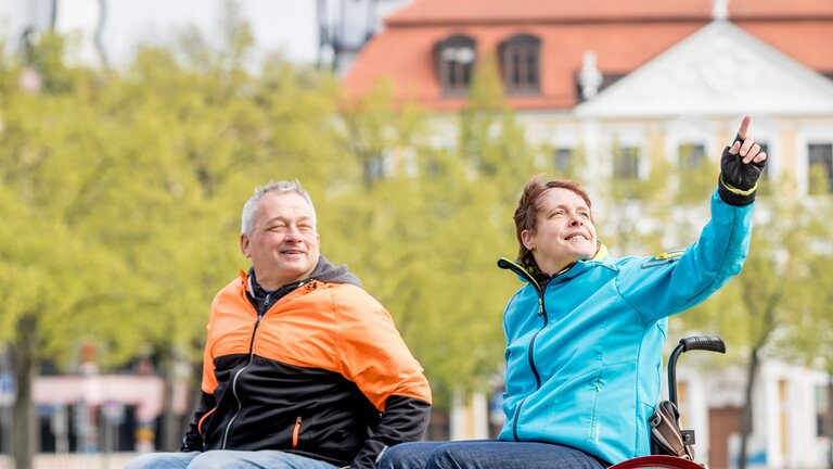 Ein Mann mit orange-schwarzer Jacke und eine Dame mit hellblauer Jacke im Rollstuhl. Die Dame zeigt mit dem Finger auf etwas. | © Magdeburg Marketing, Andreas Lander
