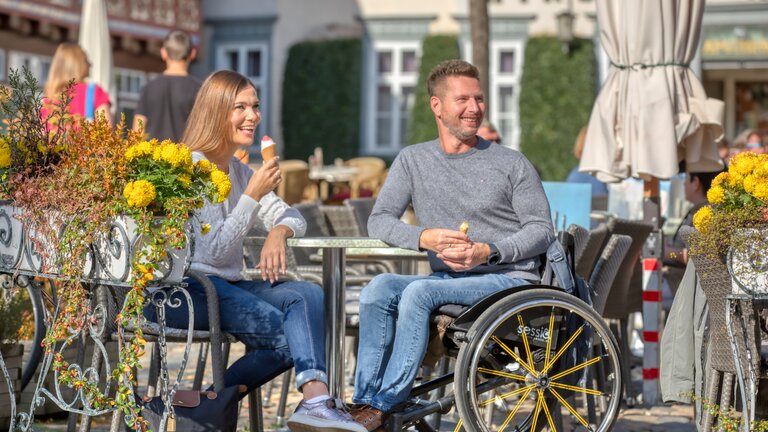 Eine junge Frau mit einem Eis in der Hand sitzt mit einem Mann in Rollstuhl an einem Tisch im Café zwischen gelben Blumen. | © Christian Bierwagen