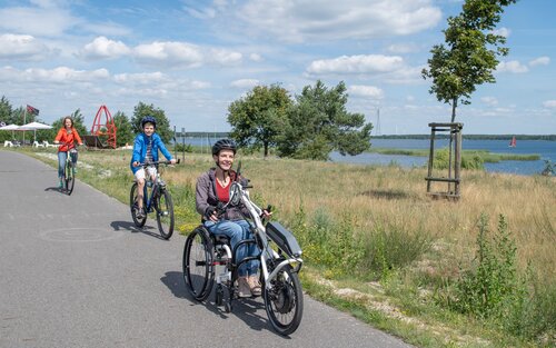 Eine Handbikerin und zwei Fahrradfahrer fahren an einem Flusslauf entlang. | © TMB-Fotoarchiv/ Nada Quenzel