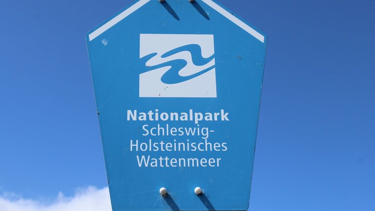 blaues Schild vor blauem Himmel mit weißer Aufschrift: „Nationalpark Schleswig-Holsteinisches Wattenmeer“ | © Frauke Feind / Pixabay