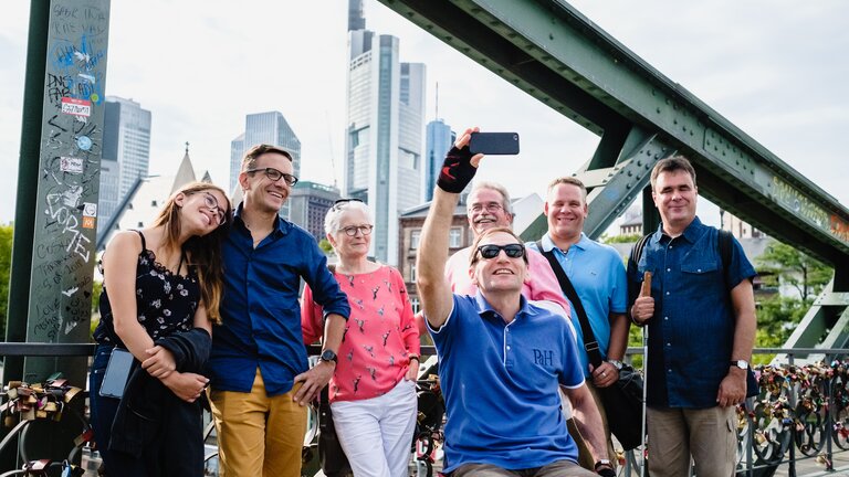 Eine Person im Rollstuhl macht ein Selfi einer Gruppe auf dem Eiserner Steg.  | © Visitfrankfurt / Andi Weiland